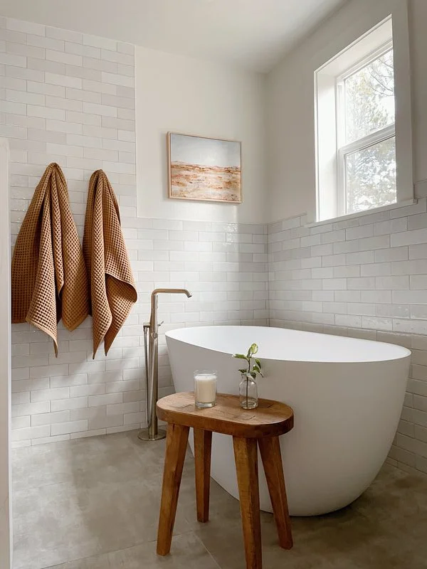 Modern bathroom with a white oval soaking tub, a small wooden stool with a candle and plant, two beige towels hanging on the wall, a window with natural light, and minimalist decor.