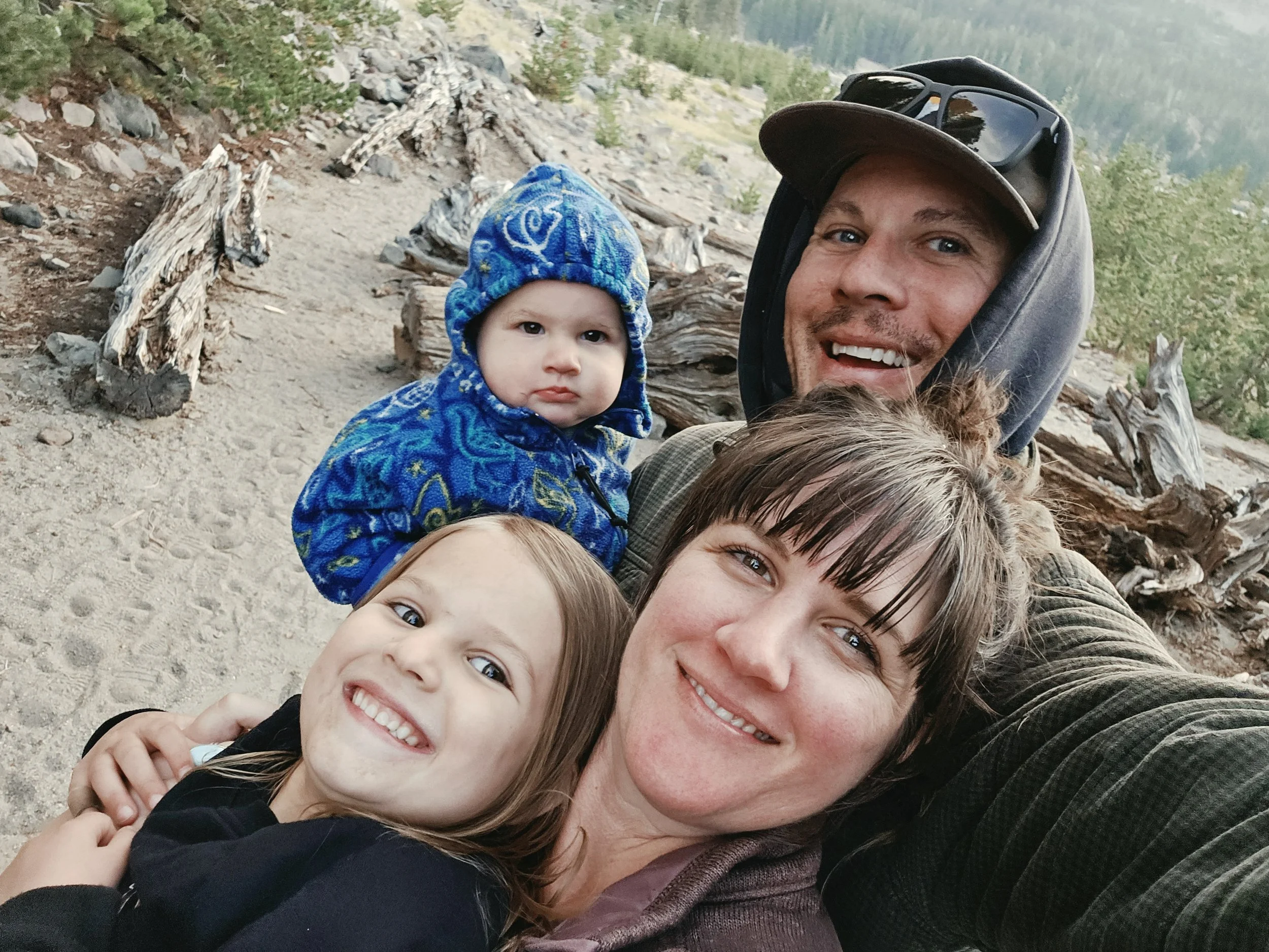 A family taking a selfie outdoors on a mountain trail with trees and rocks in the background. The group includes a man, woman, and three children, all smiling and dressed in outdoor clothing.