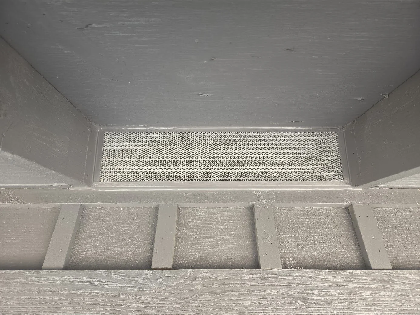 Close-up of a vent screen on a gray painted wooden porch, showing the underside and surrounding structure. Eave vents, foundation vents, roof vents, deck screening.