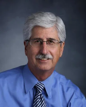 Portrait of an older man with gray hair, glasses, and a mustache, wearing a blue dress shirt and striped tie, against a gray background.