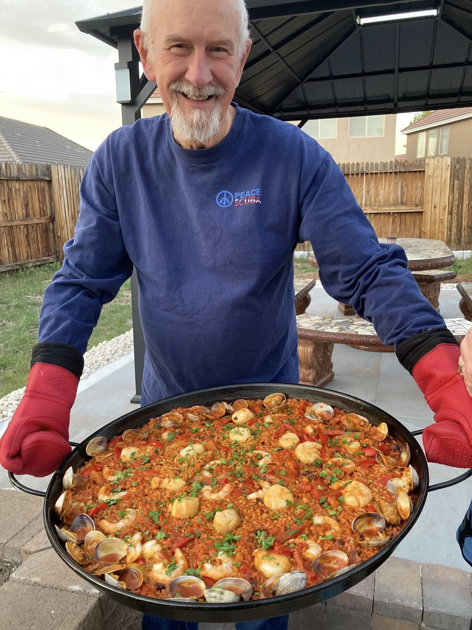 A man with white hair and a beard smiling and wearing a blue shirt with 'Peace Scuba' logo, holding a large tray of seafood paella outdoors at a backyard gathering.