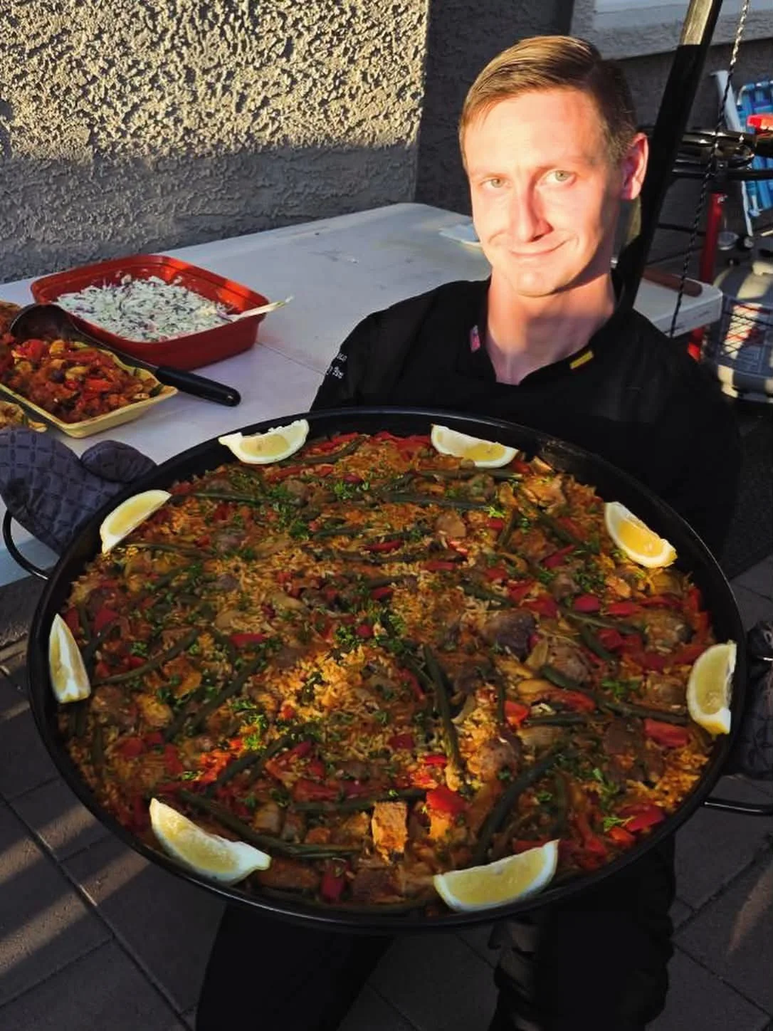 A man holding a large paella dish with lemon wedges on the edges, sitting at an outdoor table.