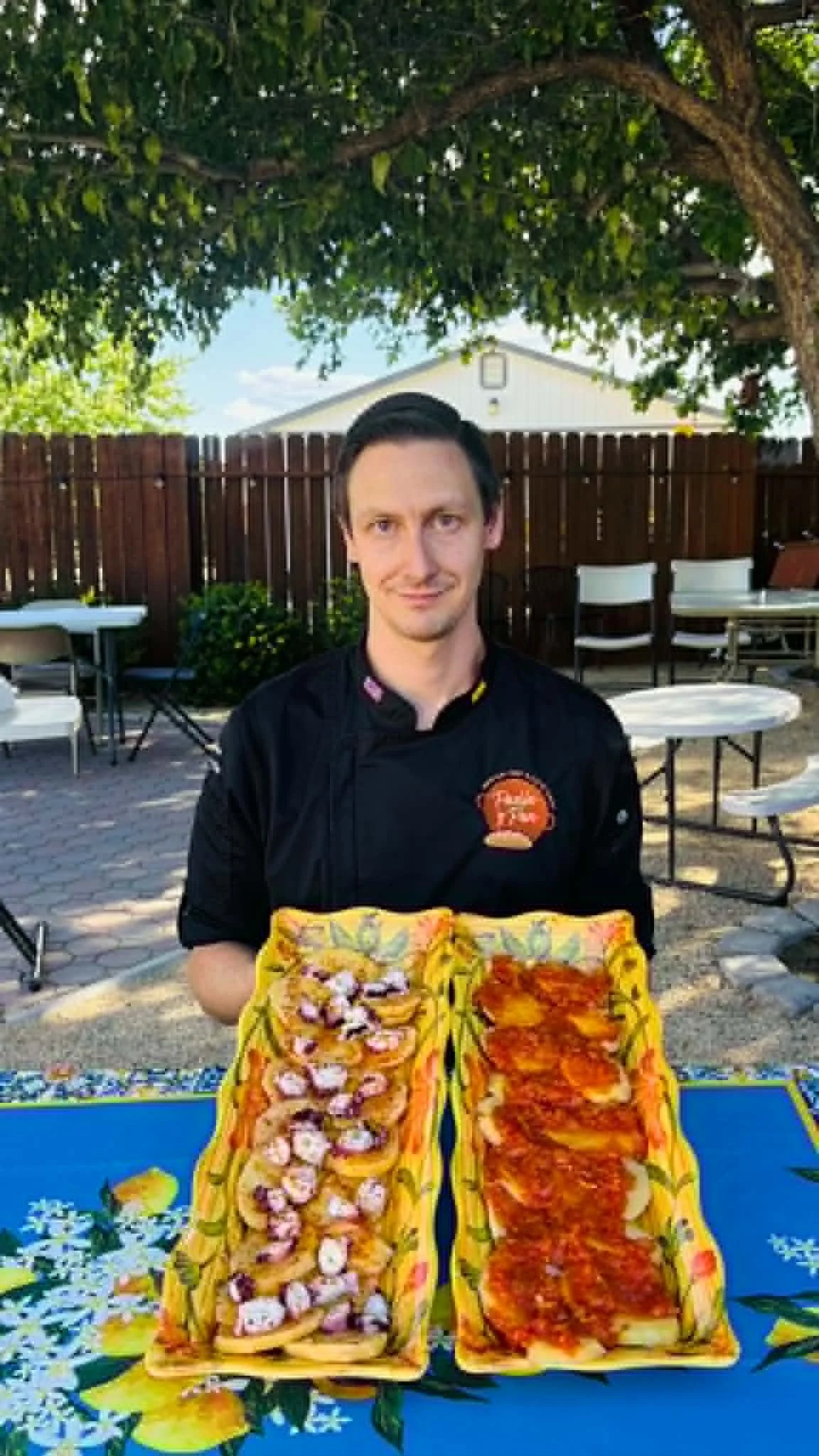 A man wearing a black chef's jacket with a logo on the chest, holding two large trays of pizza outdoors under a tree, with a wooden fence and some tables and chairs in the background.
