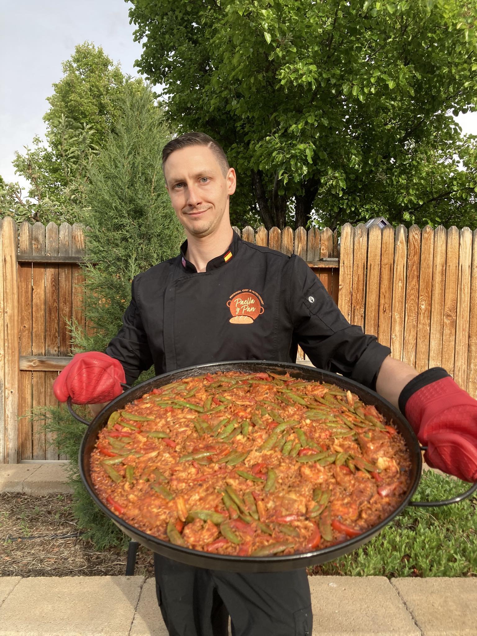 A man in a black chef's coat holding a large dish of cooked paella outdoors. The background shows a wooden fence and green trees.