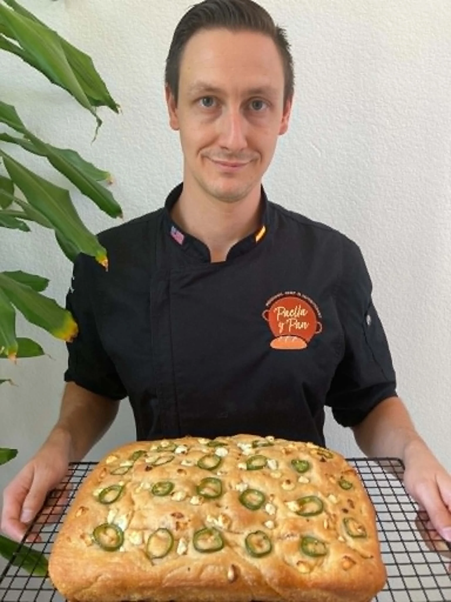 A man in a black chef's jacket with a logo, standing behind a cooling rack with a jalapeño pepper foccacia, indoors with a white wall background and a large green plant on the left.