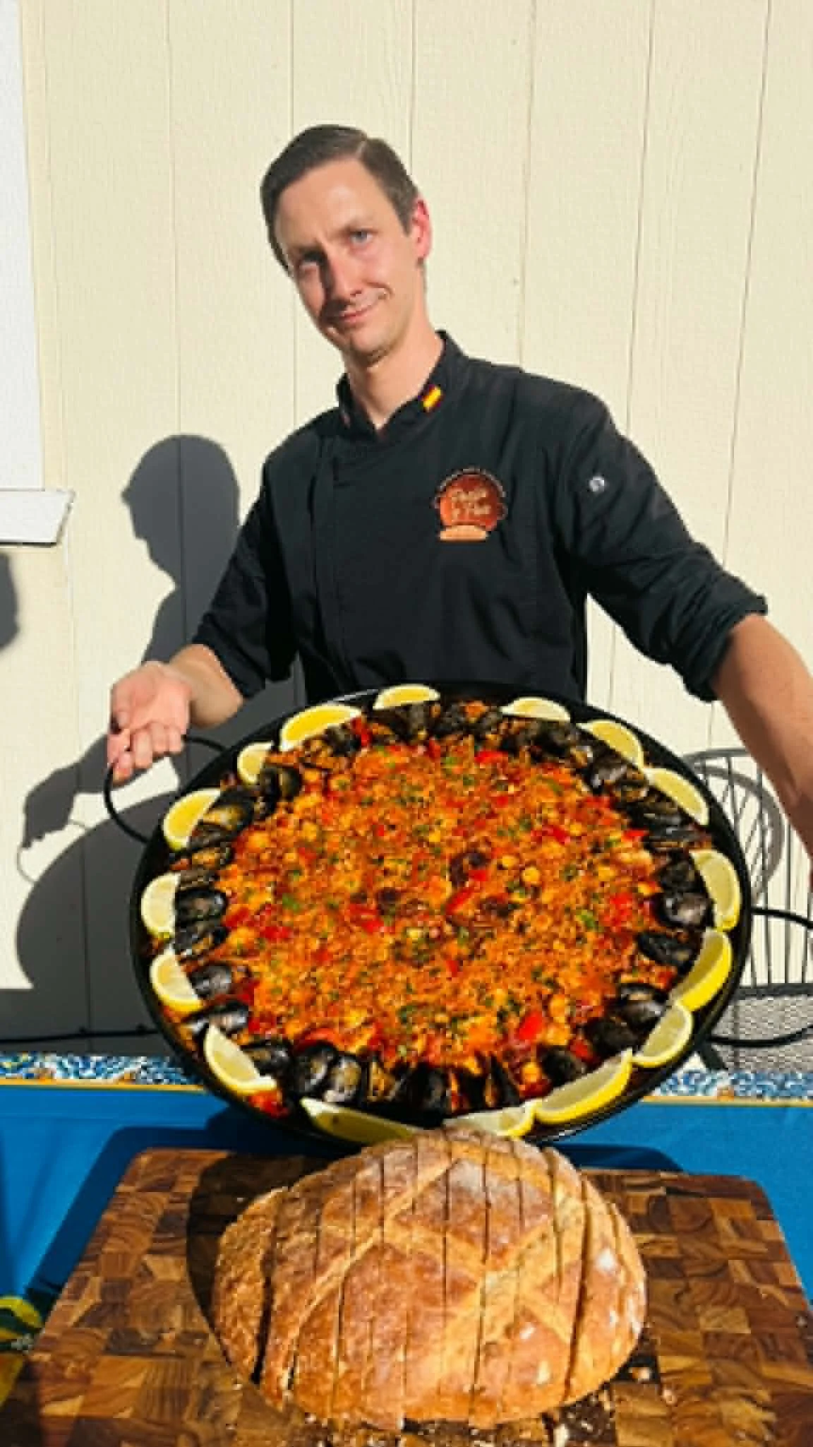 A man holding a large tray of seafood paella with lemon wedges, standing behind a table with a loaf of bread.