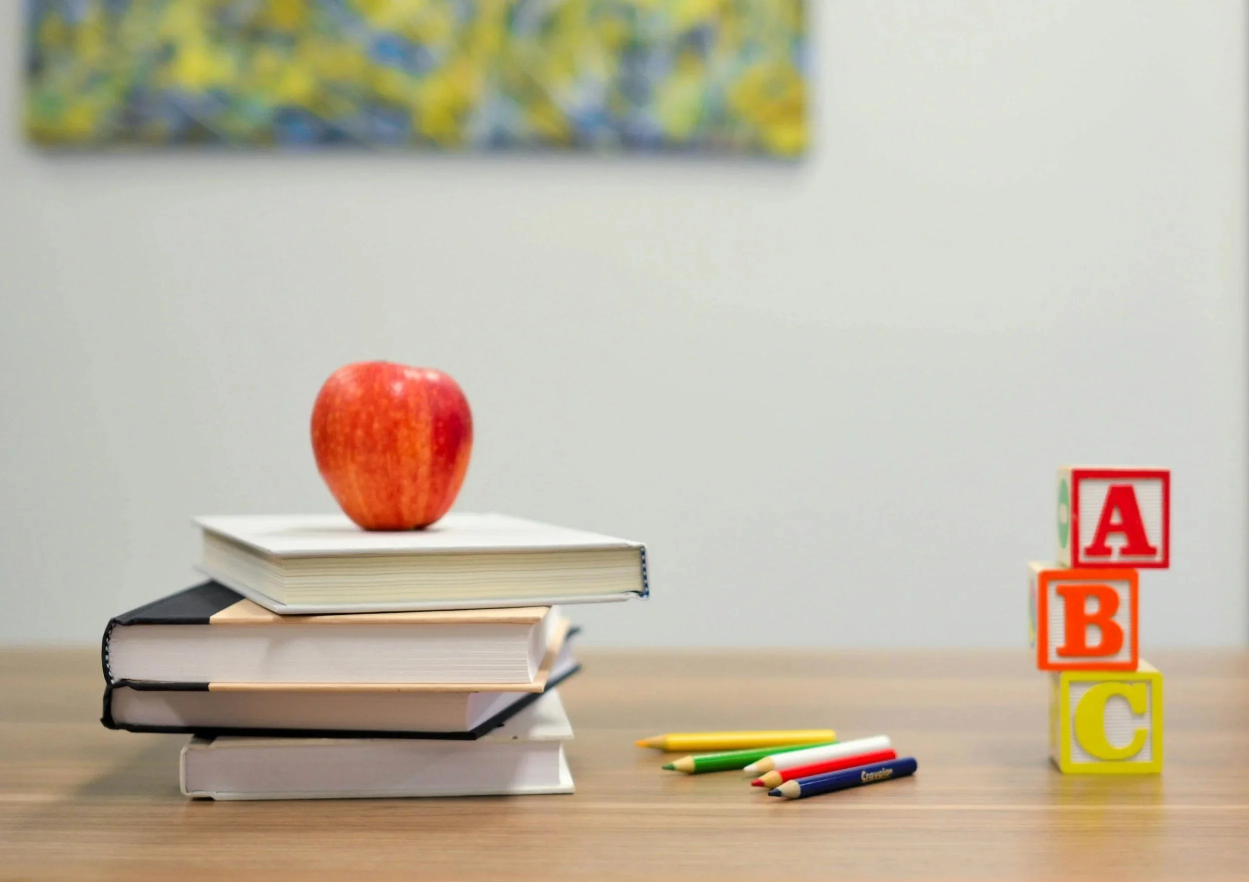 A red apple on top of a stack of books on a wooden table, with colorful alphabet blocks and pens nearby, set against a plain wall with a blurred colorful picture hanging.