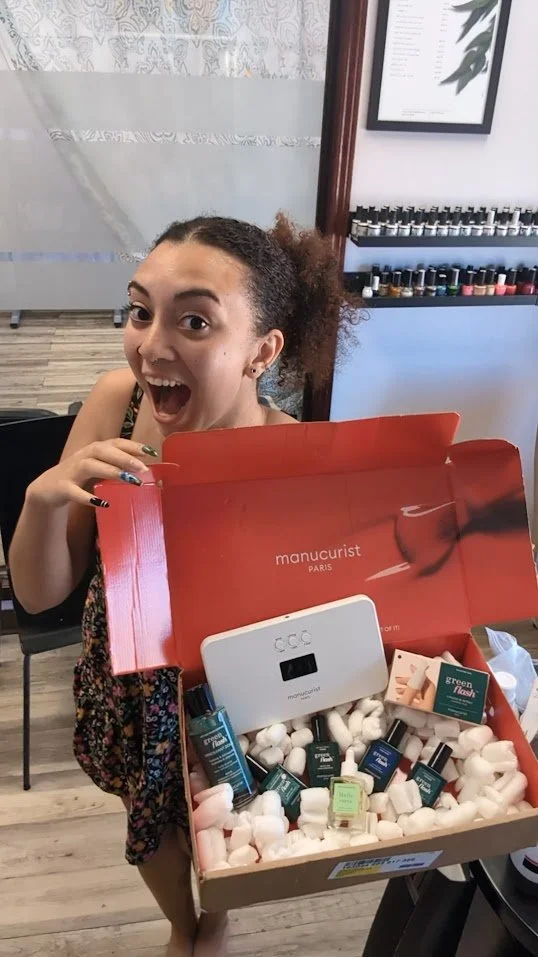 A woman with curly hair holding a Manucurist Paris nail care product box, smiling excitedly inside a nail salon.
