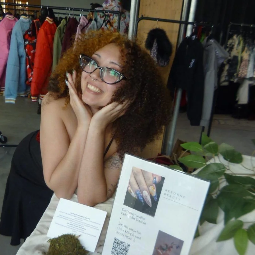 A woman with curly two-tone hair, wearing glasses, is smiling and posing with her hands on her cheeks in a clothing store. There are racks of colorful jackets and coats behind her.