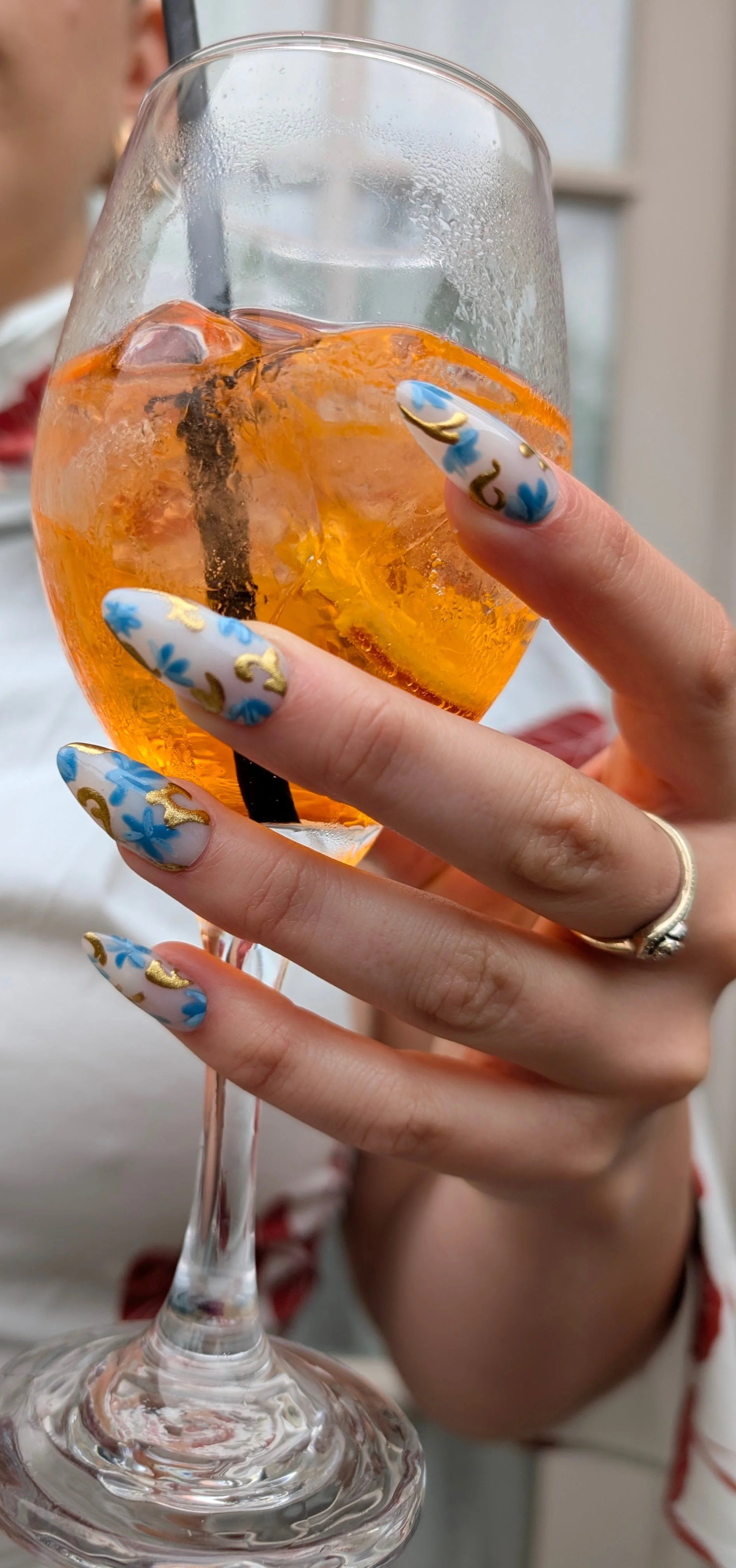 A person's hand holding a wine glass filled with an orange beverage and ice, with a blurred background.