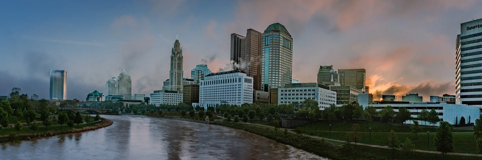 Skyline of a city with tall buildings and a river in the foreground during sunrise.