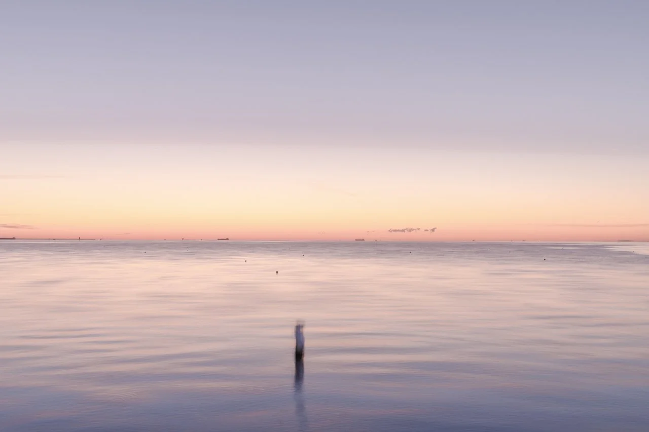 A calm ocean scene during sunset with a colorful sky reflecting on the water, and a single weathered post sticking out of the water in the foreground.