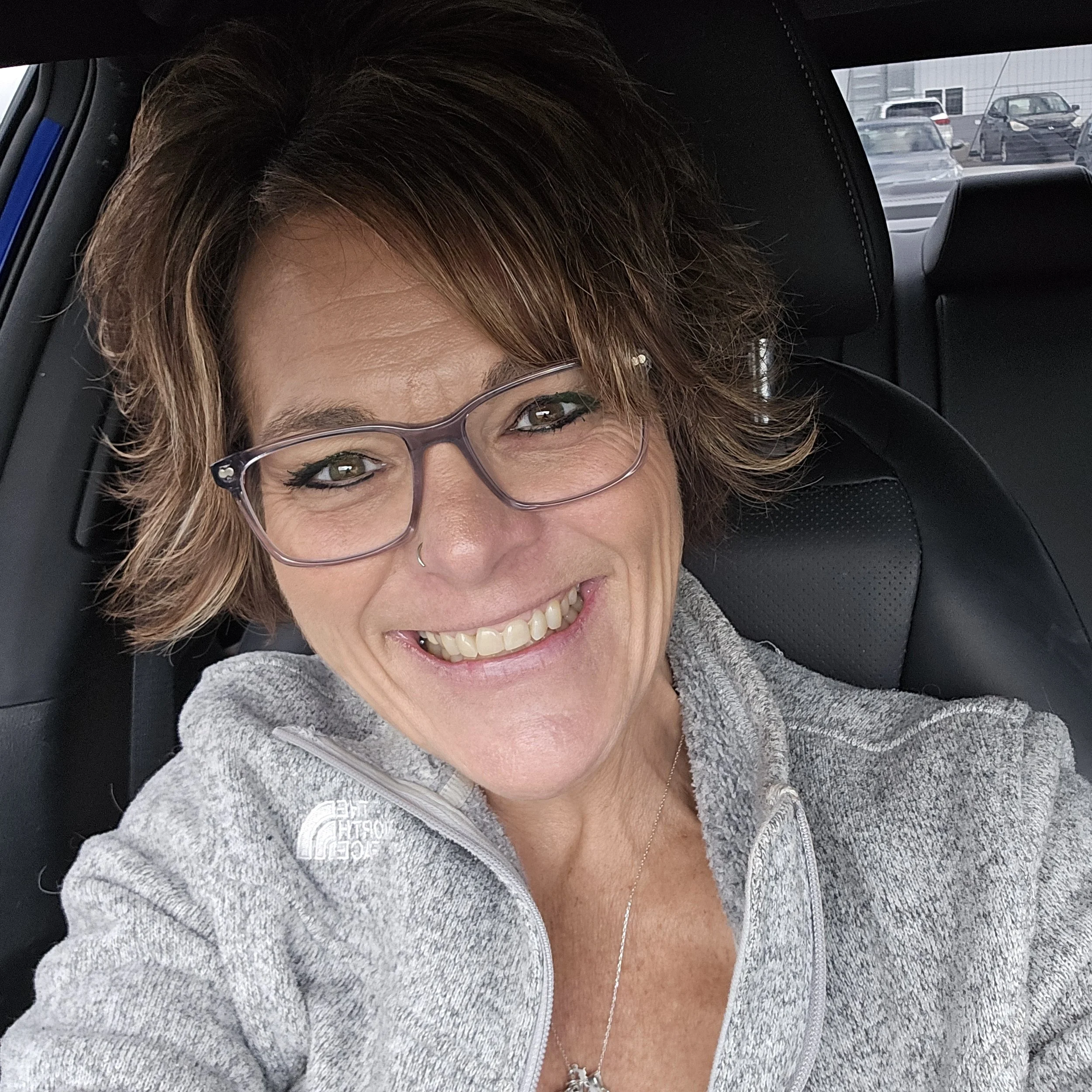 A woman with brown hair and glasses smiling in a car, wearing a gray zip-up fleece and a necklace.