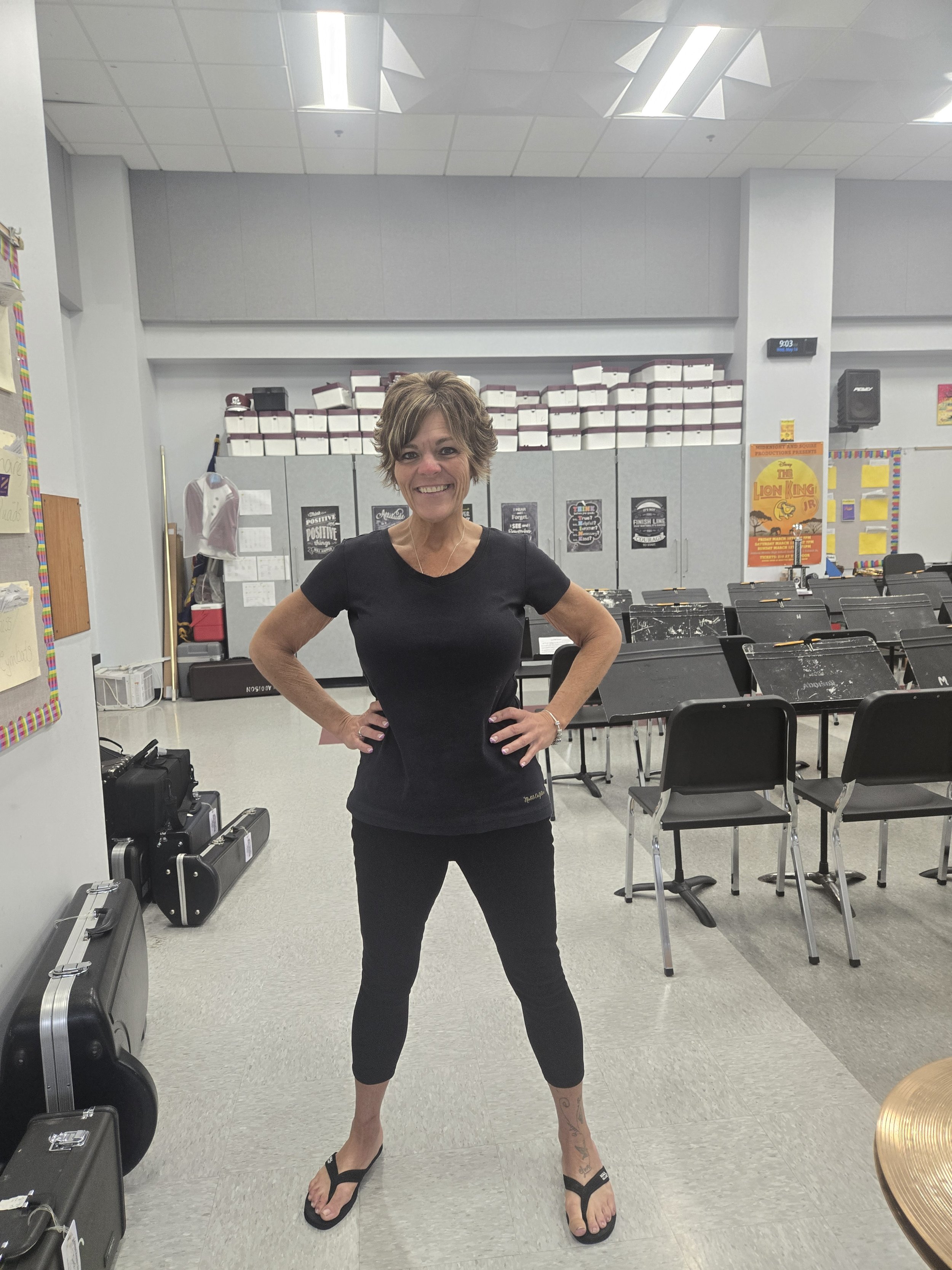 A woman smiling and posing with hands on hips inside a classroom or rehearsal space, with music stands, chairs, posters, and equipment visible in the background.