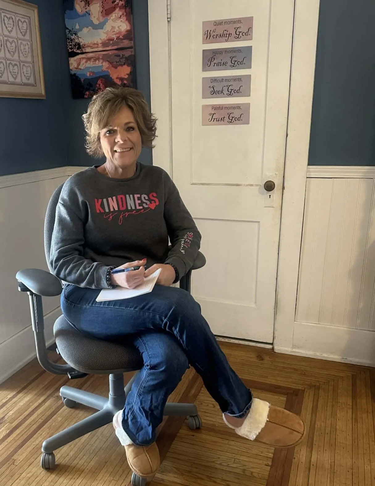 A woman sitting in a rolling office chair, holding a notepad and pen, smiling at the camera. She is wearing a sweatshirt with the words 'KINDNESS is free', jeans, and slippers. Behind her is a white door with signs about worship and gratitude, and artwork on the walls.