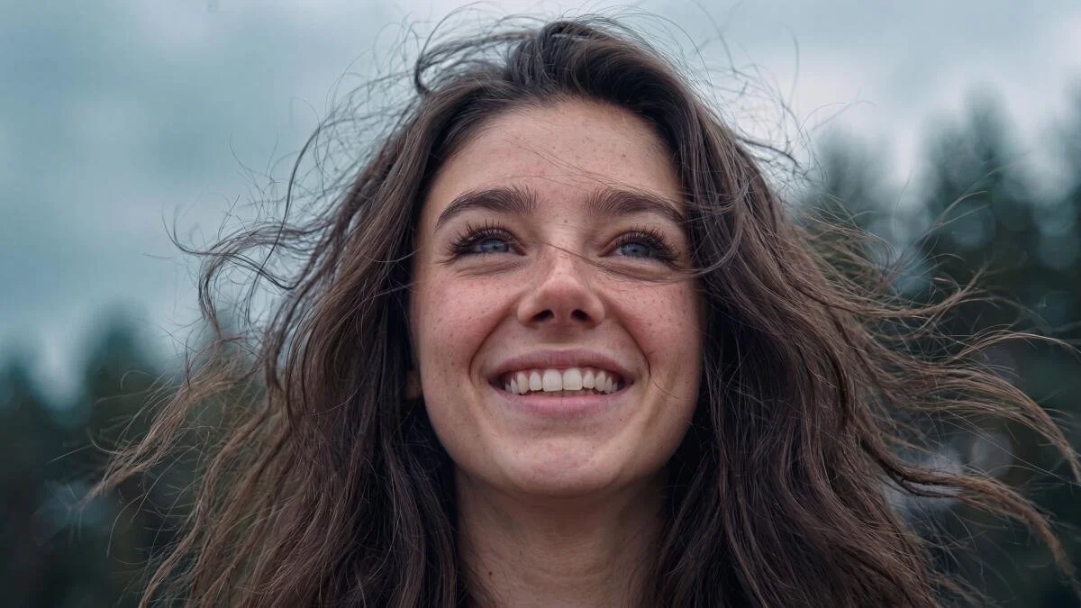 Close-up of a young woman smiling outdoors, with her hair blowing in the wind.