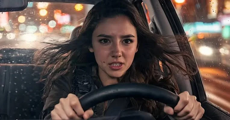 A young woman with long brown hair driving a car at night, showing a tense or angry expression.