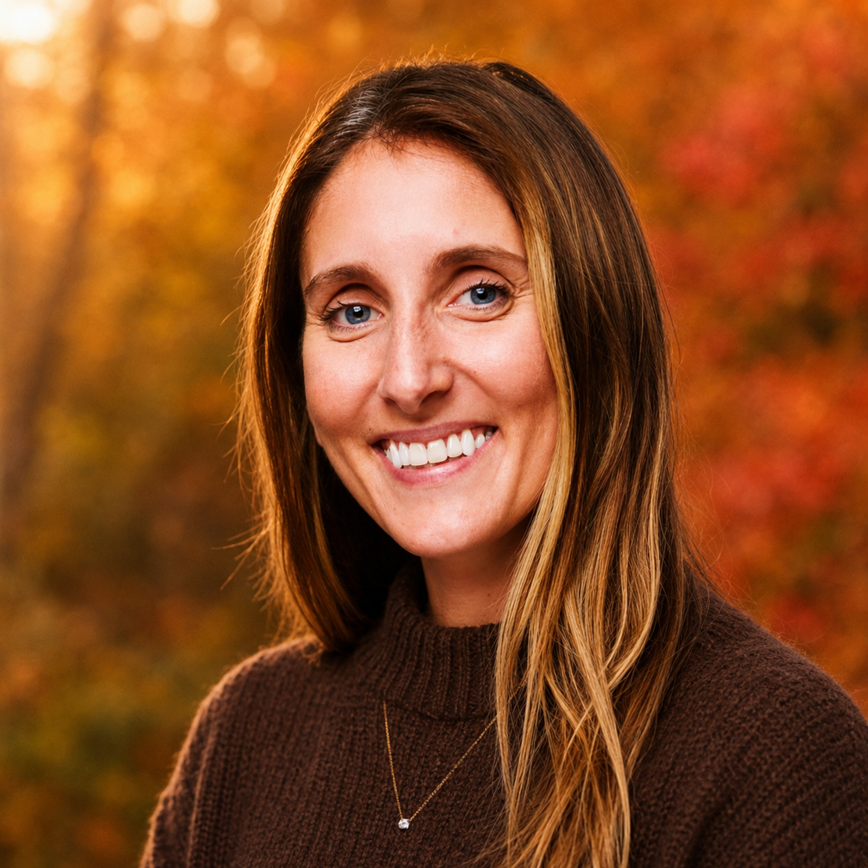 A smiling woman with long brown hair and blue eyes outdoors during fall, wearing a dark brown sweater and a small necklace.