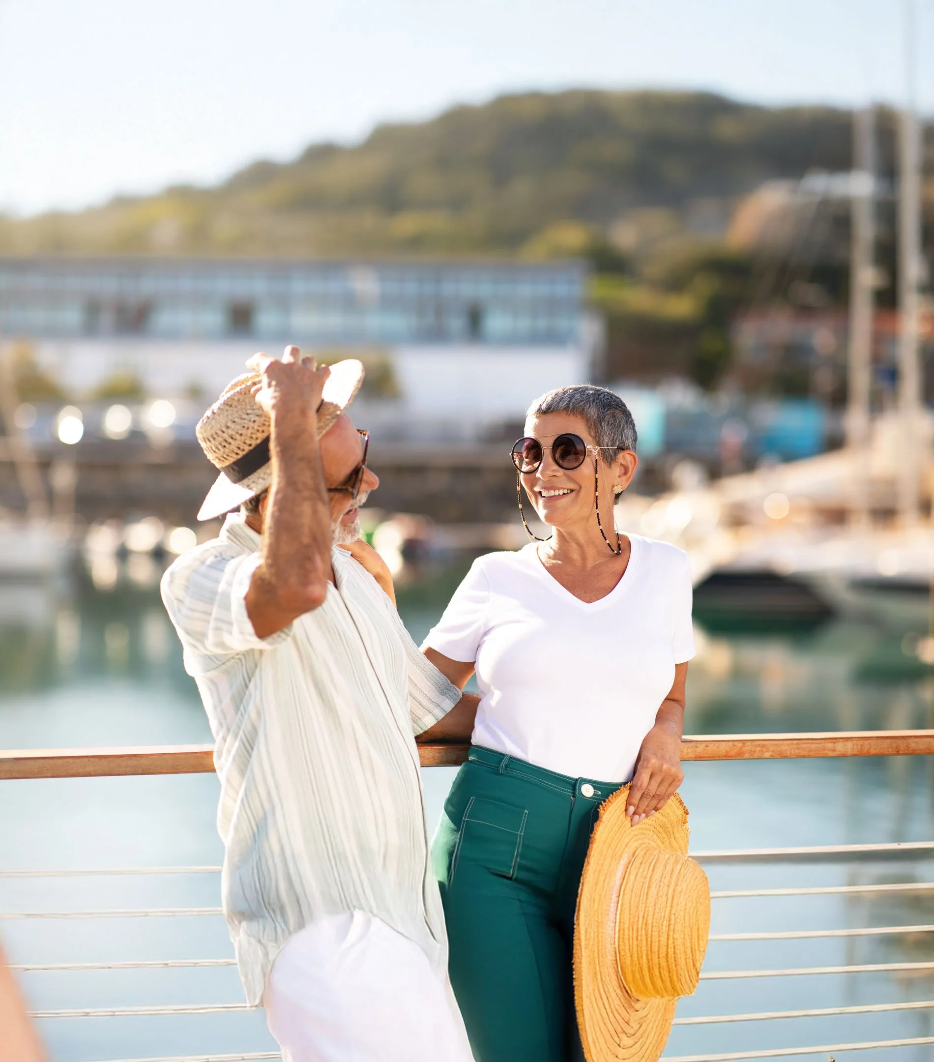 Two older women standing near a body of water with boats, wearing sunglasses and smiling at each other, one holding a large straw hat and a yellow woven bag, with a green hill and a bridge in the background.
