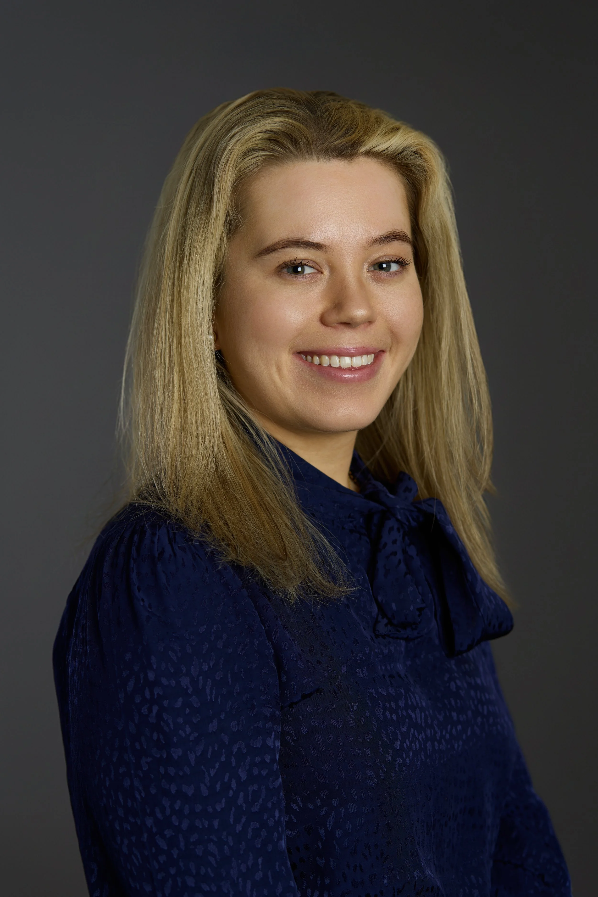 A woman's close-up portrait with blonde hair, smiling, wearing a dark blue textured blouse, against a dark gray background.