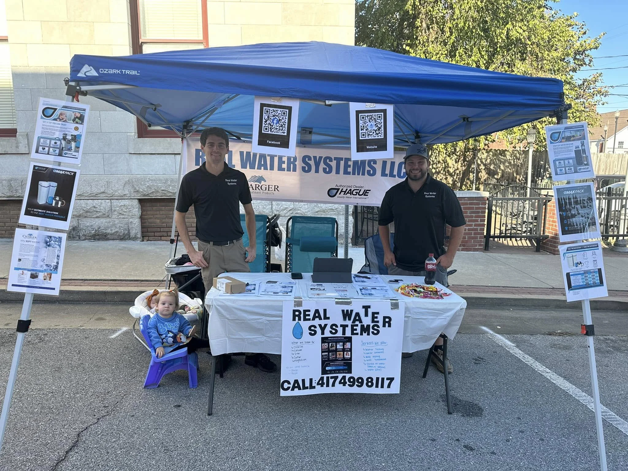 A booth at an outdoor event promoting real water systems, manned by two men in black shirts smiling at the camera. The booth has a table with informational materials and a banner displaying the company's contact details. QR codes promote Facebook and the company website. There are posters on display, and a small child in a purple chair with a feeding bottle is in the foreground.