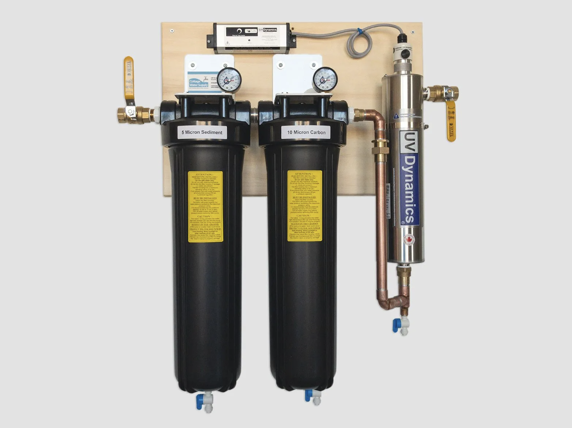 Water filtration system mounted on a wooden panel with two black filter tanks labeled '5 Micron Sediment' and '10 Micron Carbon,' alongside a cylindrical UV filter labeled 'UV Dynamics' connected with copper piping and valves.