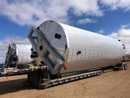 Large cylindrical industrial tank being transported on a flatbed trailer in an open area with a partly cloudy sky.