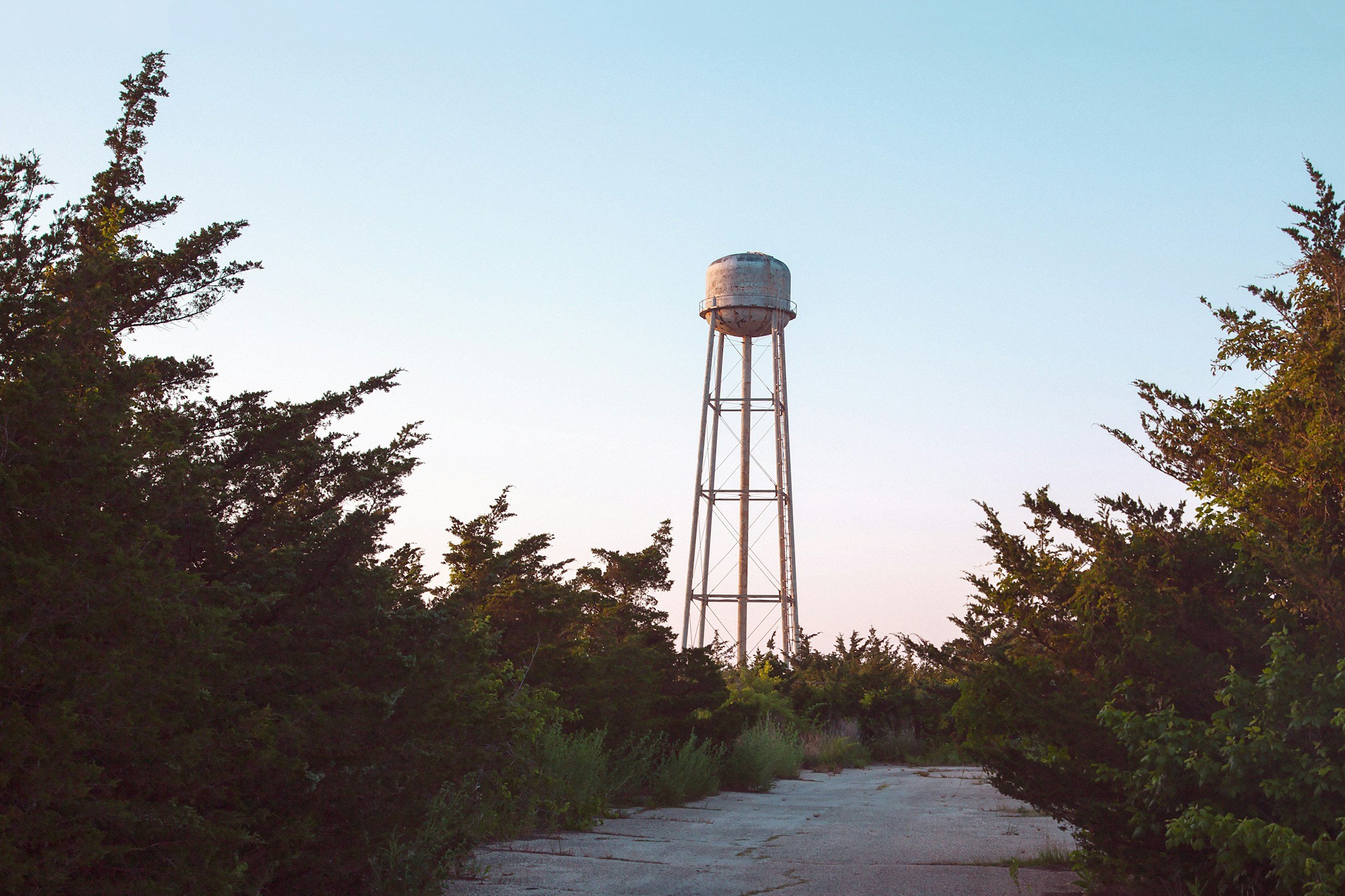 A rusted water tower stands tall amidst green trees under a clear sky.