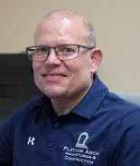 A man with glasses and a shaved head wearing a blue collared shirt.