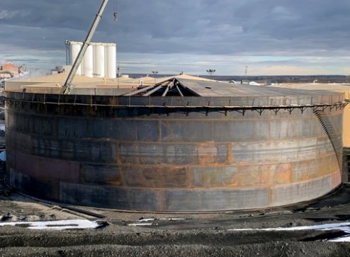 A large, circular industrial storage tank with rusted metal exterior and a damaged roof, located outdoors against a cloudy sky.