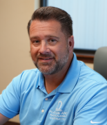 A man with short, dark hair and a beard, wearing a light blue collared shirt, sitting in an office environment.