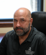 A middle-aged man with a bald head and beard sitting at a desk in an office.