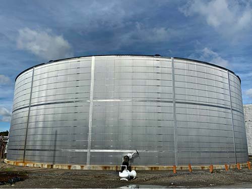 Large cylindrical industrial storage tank made of metal with a vent or pipe at the bottom, situated outdoors under a cloudy sky.