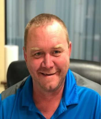 A man with short light brown hair and a goatee, smiling, wearing a blue polo shirt, sitting in an office with vertical blinds in the background.