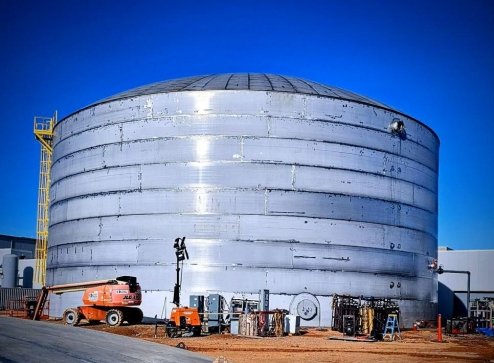 Large silver industrial storage tank with equipment and machinery nearby, under a clear blue sky.