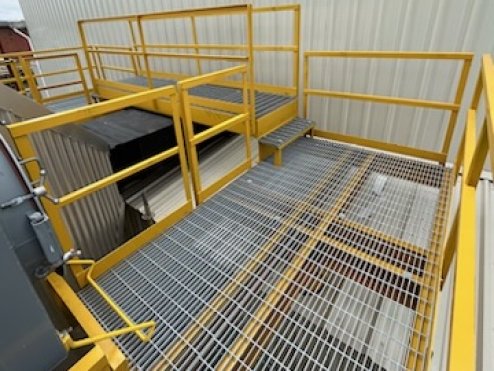 Yellow metal safety platform with grated floors and guardrails in an industrial setting.