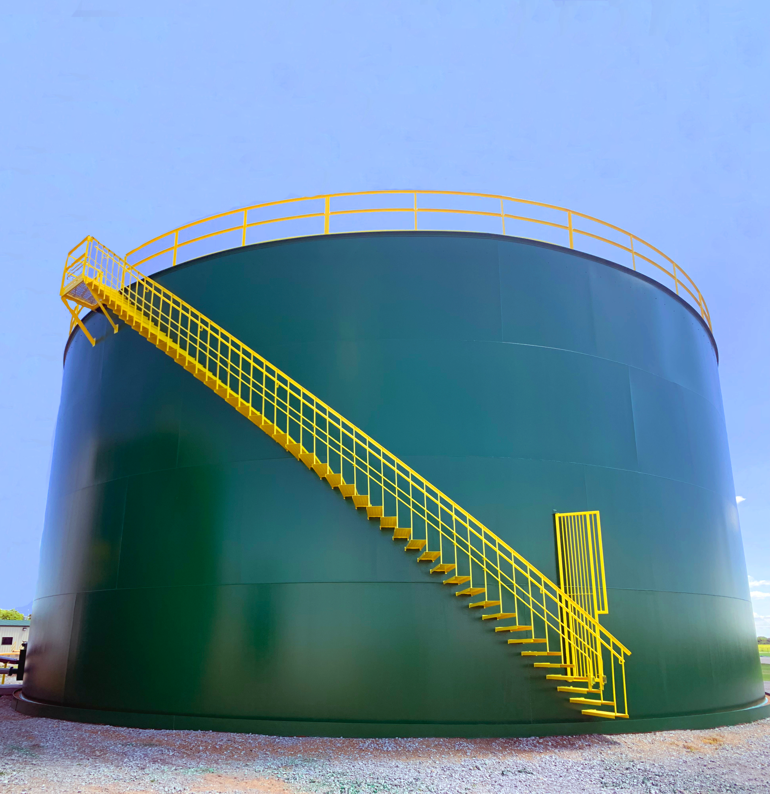A large green industrial storage tank with yellow safety railings and stairs, set against a clear blue sky.