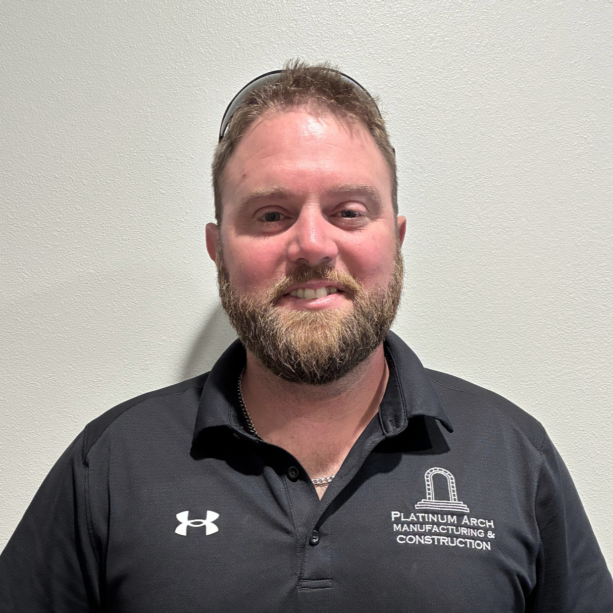 A man with a beard and short hair, wearing sunglasses on his head and a black collared shirt with the logo and text 'Platinum Arch Manufacturing & Construction.' standing against a plain white wall.