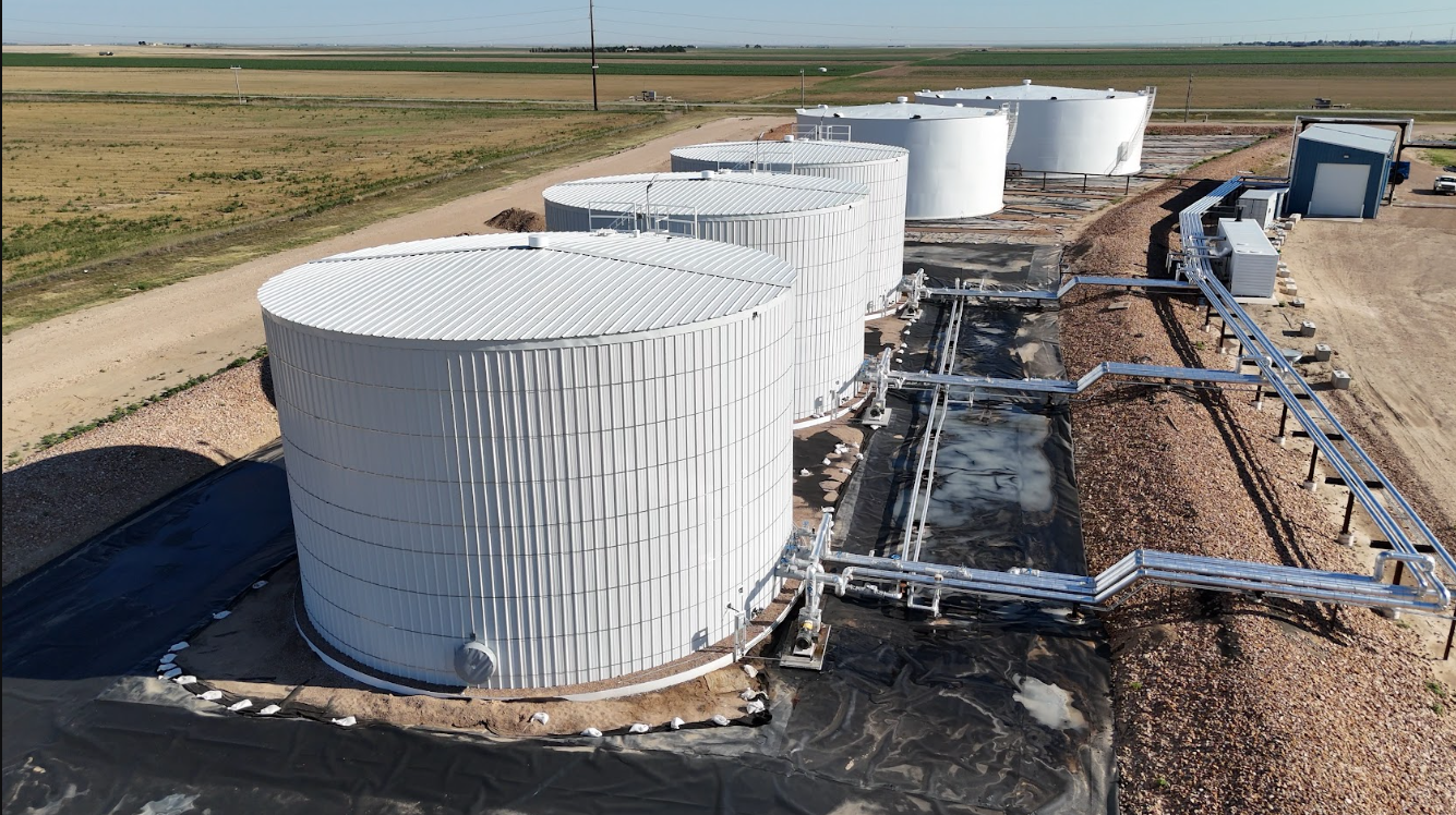 Large white industrial storage tanks connected by piping, situated in a flat, open landscape, likely part of a facility such as a liquid storage or processing plant.