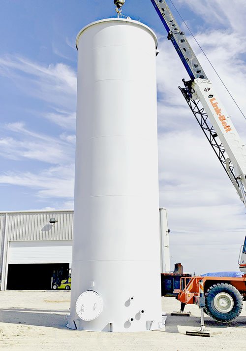 A tall white cylindrical industrial storage tank being lifted by a crane outside a warehouse building.