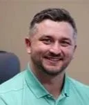 Headshot of a man with dark hair, beard, and mustache, smiling, wearing a light green shirt, against a gray background.