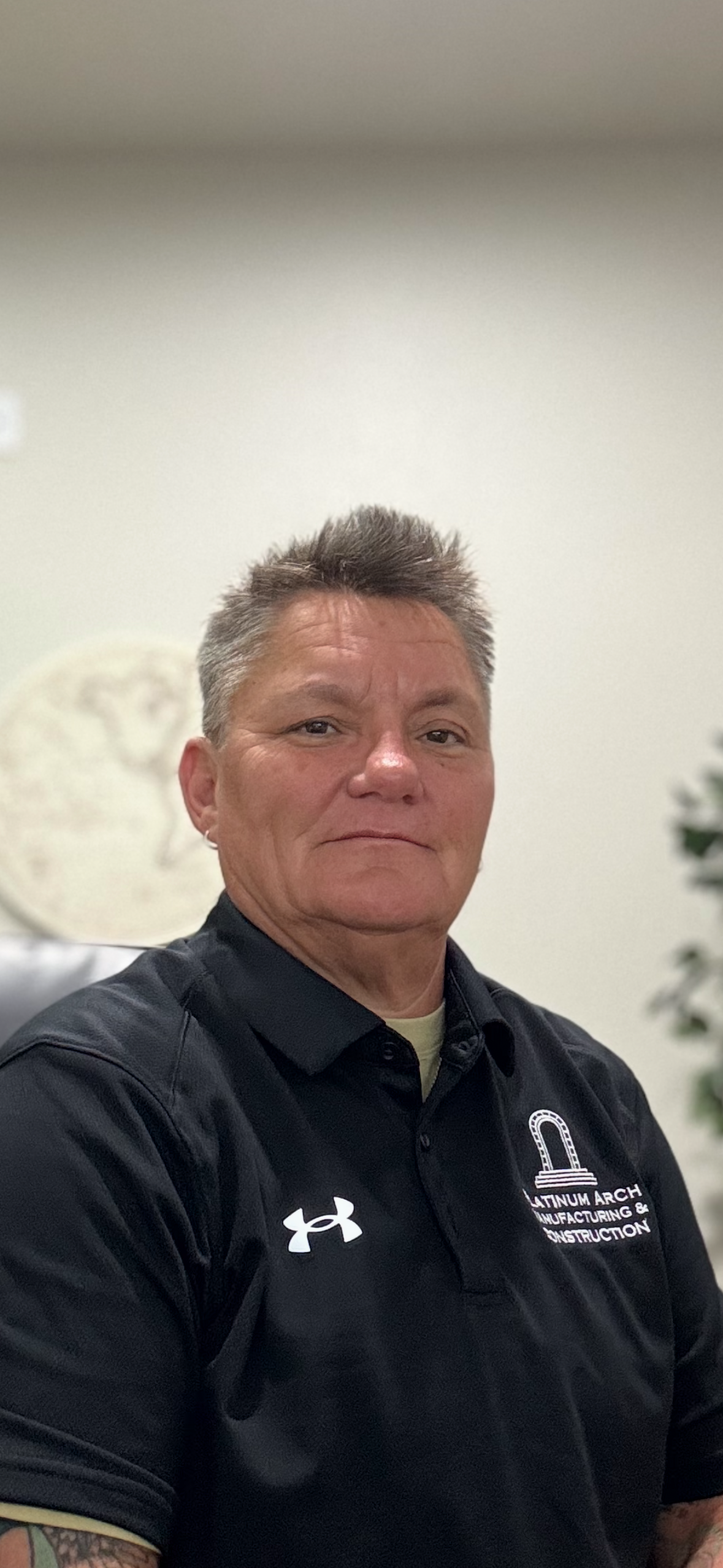 A woman with short gray hair is taking a selfie in an office or lobby. She is wearing a black collared shirt with a logo that reads 'Platinum Arch Manufacturing & Construction' and the Under Armour logo. 