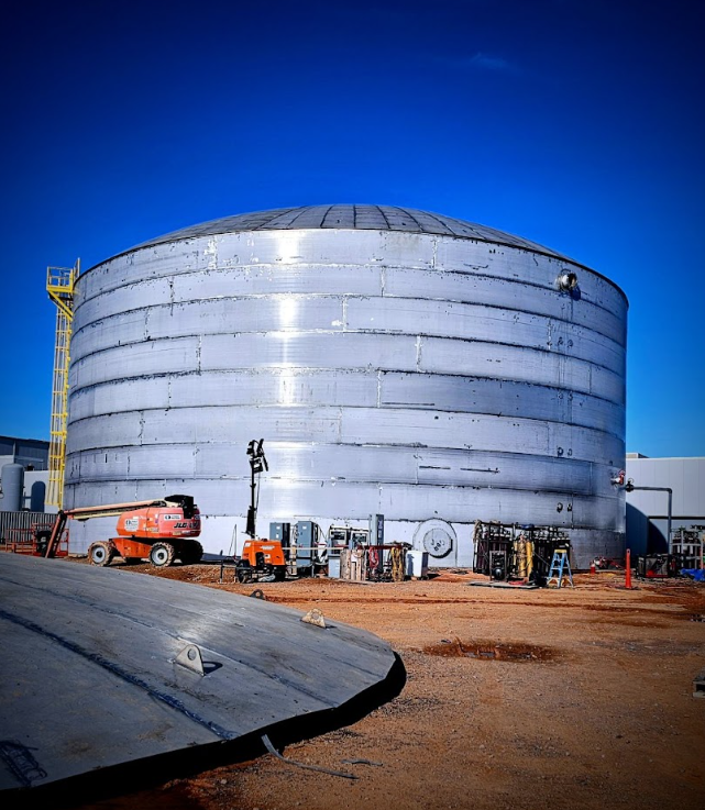 Large metallic cylindrical storage tank at an industrial site with construction equipment nearby.
