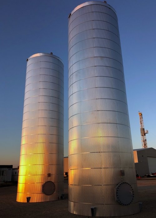Two tall, cylindrical metallic grain silos standing outdoors against a clear evening sky, illuminated by yellow-orange lighting at the base.