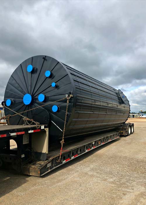 A large black cylindrical industrial tank securely fastened on a flatbed trailer in an open area with cloudy skies overhead.