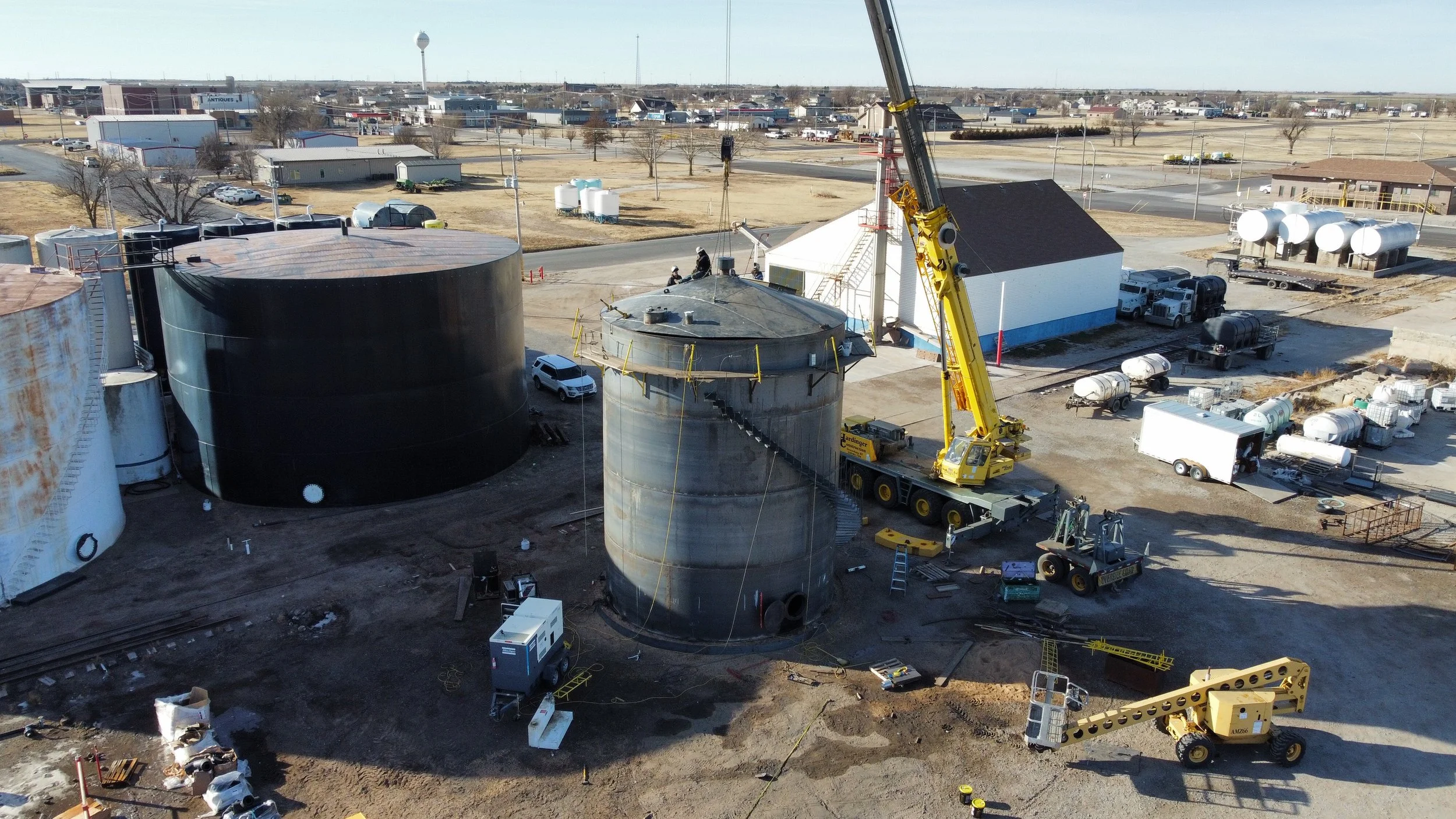 Construction site with large cylindrical tanks, a crane lifting equipment, and workers on the roof of a small building. Background shows a semi-urban area with trees, roads, and scattered buildings.