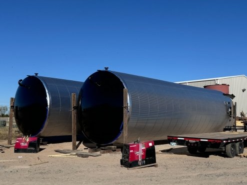 Two large black cylindrical tanks outdoors on a dirt surface, with a clear blue sky and industrial building in the background.