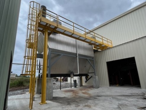 Industrial structure with a yellow catwalk and ladder, connected to a large metallic tank or container next to a warehouse building, on a cloudy day.
