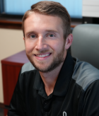 A man with short brown hair and a beard, smiling, sitting in an office chair in front of a desk with a filing cabinet.