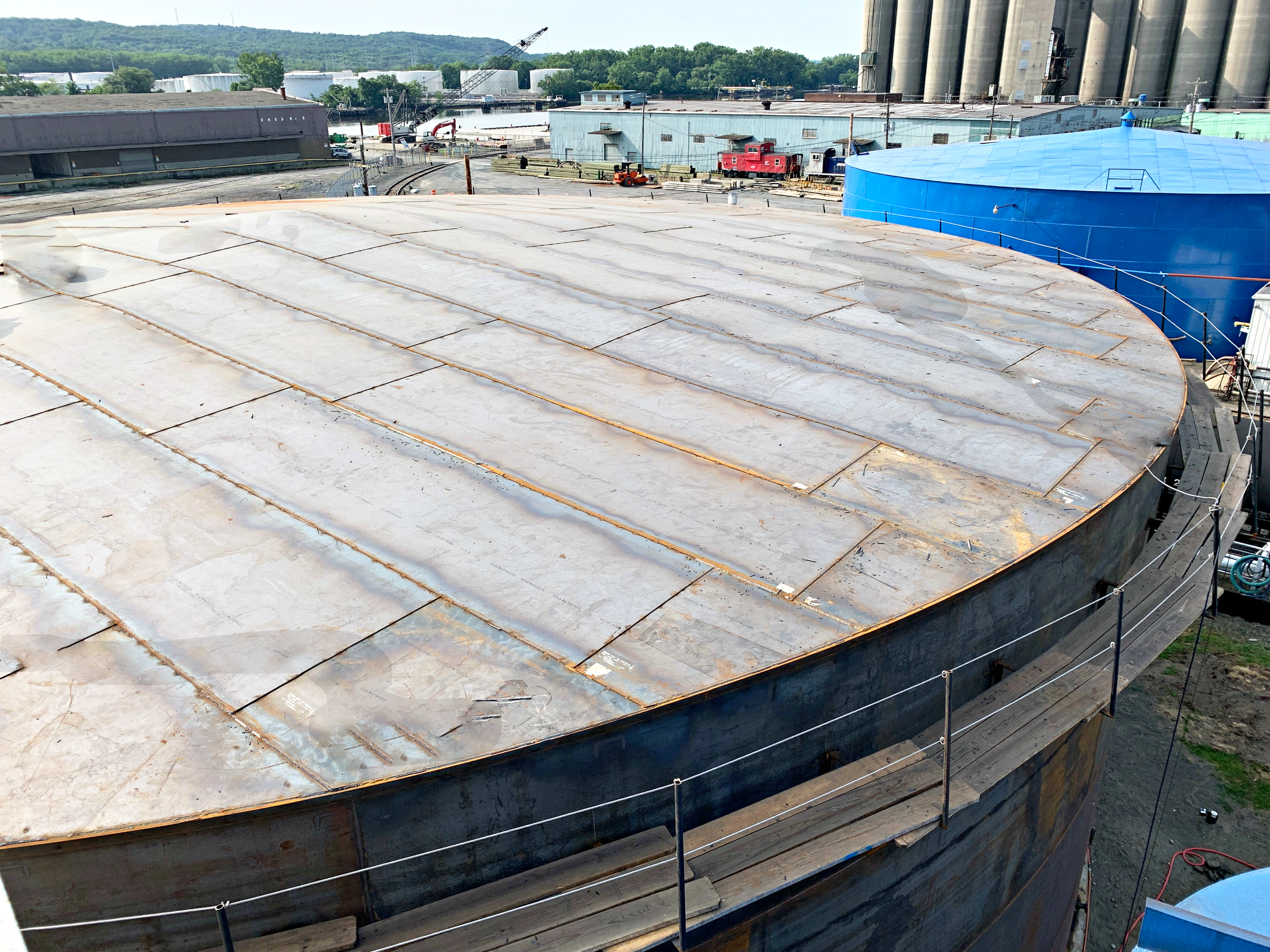 Construction site with a large curved metal roof on storage tank, industrial buildings, and silos in the background. Various construction materials and equipment are visible.